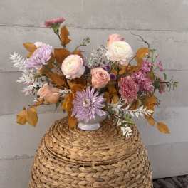 Pastel pink and peach floral arrangement with lavender mums and autumn leaves in a white vase on a woven basket.