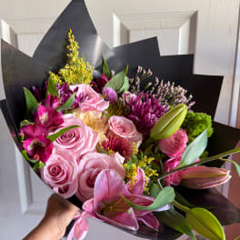 Handheld bouquet of pink roses, lilies, and purple flowers wrapped in black paper