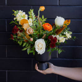 Autumn-toned bouquet of roses and ranunculus in a black pedestal vase held against a dark wall