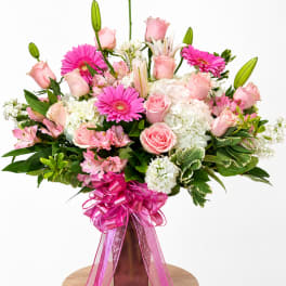 Pink roses, gerbera daisies, lilies, and white hydrangeas in a tall vase with a pink ribbon