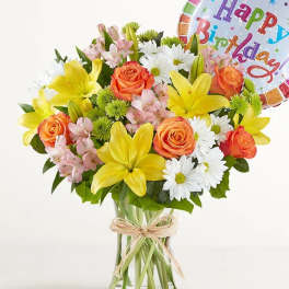 Bouquet of yellow lilies, orange roses, and white daisies in a glass vase with a birthday balloon