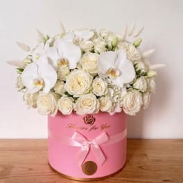 White roses and orchids arranged in a pink round box with a ribbon.