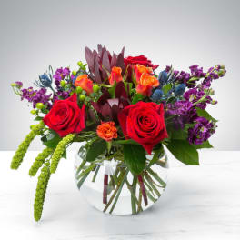 Mixed bouquet of red roses, orange blooms, and purple flowers in a glass vase