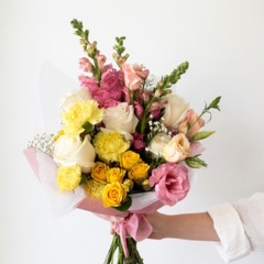Handheld bouquet of pink, yellow, and white flowers wrapped in pale pink paper