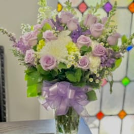 Lavender rose bouquet with white blooms in a clear glass vase and purple ribbon