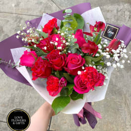 Hand-tied bouquet of red roses, carnations, and white filler flowers wrapped in purple and white paper.