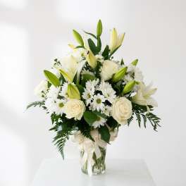 White lilies, roses, and daisies arranged in a glass vase with a ribbon