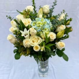 Tall glass vase of cream roses, white lilies, and blue hydrangea with mixed filler flowers.