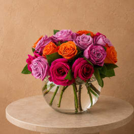 Mixed roses in pink, purple, and orange arranged in a clear glass bowl vase