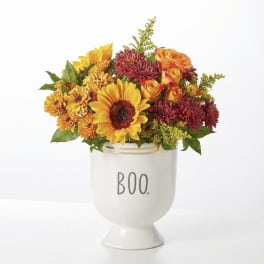 Mixed orange, yellow, and burgundy flowers in a white vase labeled BOO.