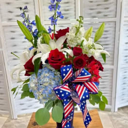 Red roses, white lilies, blue flowers, and a patriotic ribbon in a vase