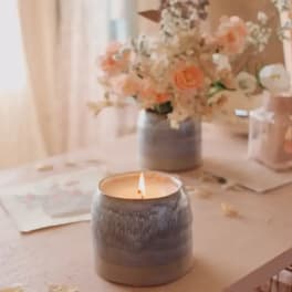 Lit candle in a ceramic holder with a peach floral arrangement in the background