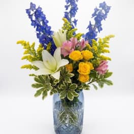 Blue and yellow mixed bouquet in a glass vase with white lilies and pink flowers