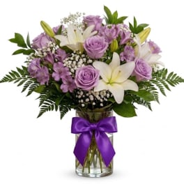 Lavender roses and white lilies in a glass vase with a purple ribbon