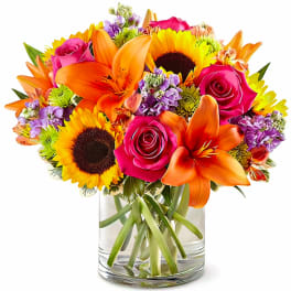 Bright mixed bouquet of sunflowers, roses, lilies, and purple flowers in a glass vase