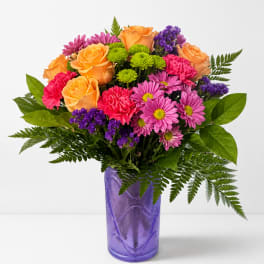Mixed bouquet of orange roses, pink daisies, and purple filler in a purple vase
