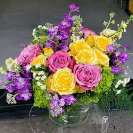 Arrangement of pink and yellow roses with purple stock and green hydrangeas in a clear glass vase.