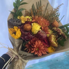 Hand-tied bouquet of orange mums and yellow roses wrapped in kraft paper with a raffia bow