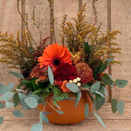 Autumn arrangement of orange gerbera, roses, and mums in a pumpkin container with eucalyptus accents