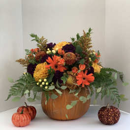 Autumn bouquet of orange and yellow flowers in a pumpkin container with small decorative pumpkins nearby.