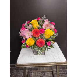 Bright mixed bouquet with yellow roses, pink flowers, and orange blooms in a square vase
