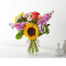 Mixed bouquet with a sunflower, rose, and pink blooms in a clear glass vase