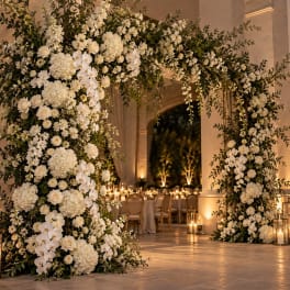 White floral arch with roses, hydrangeas, and orchids in a warm indoor venue