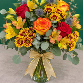 Mixed bouquet of roses, lilies, and chrysanthemums in a glass vase with a gold ribbon