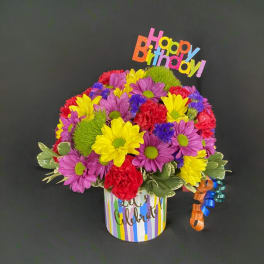 Colorful mixed flower arrangement in a striped birthday container with a Happy Birthday topper