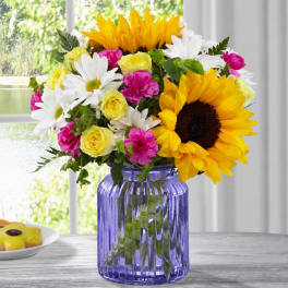 Sunflowers, white daisies, yellow roses, and pink carnations in a purple glass vase