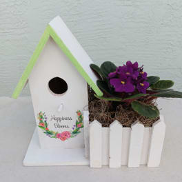 White birdhouse planter with purple flowers in a small picket fence box