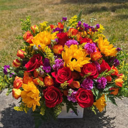 Bright mixed bouquet of red roses, yellow blooms, and purple flowers in a white container