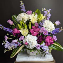 Lavender and white floral arrangement with roses, lilies, and hydrangeas in a stone vase