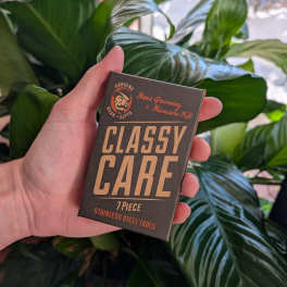 Hand holding a boxed men's grooming and manicure kit in front of large green leaves