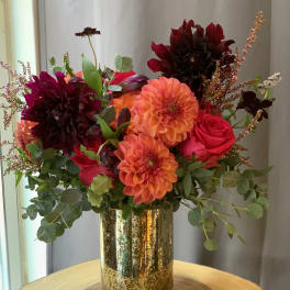 Bright mixed bouquet in a gold vase with orange, red, and dark burgundy blooms