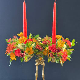 Two red taper candles rise from a colorful floral centerpiece with a skeleton base.