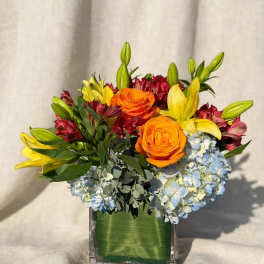 Orange roses and yellow lilies in a square glass vase with blue hydrangeas