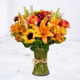 Orange lilies, roses, and sunflowers in a glass vase with a twine bow