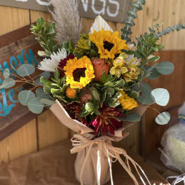 Rustic bouquet of yellow sunflowers, orange roses, and burgundy blooms wrapped in brown paper and raffia.