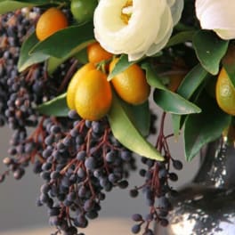 Arrangement with a white flower, orange citrus fruits, and dark berries in a dark vase