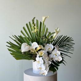 White orchids and calla lilies arranged with palm fronds in a white vase