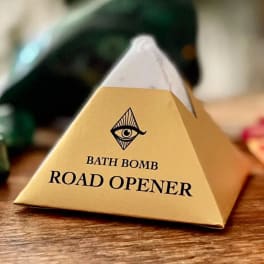 Pyramid-shaped bath bomb package labeled Road Opener on a wooden surface.