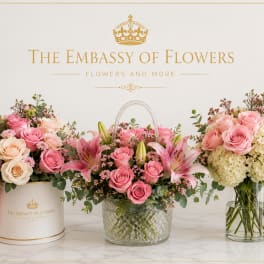 Three pink rose and lily arrangements in containers with a gold logo backdrop