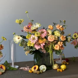 Mixed pastel flower arrangement in a green glass vase with fruit on the table