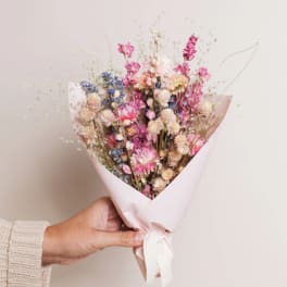 Handheld bouquet of dried pink, cream, and blue flowers wrapped in pale paper