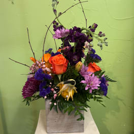 Mixed bouquet with orange roses, purple flowers, and a yellow decorative accent in a square vase