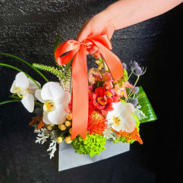 Handheld floral basket with white orchids and bright mixed blooms tied with a coral ribbon