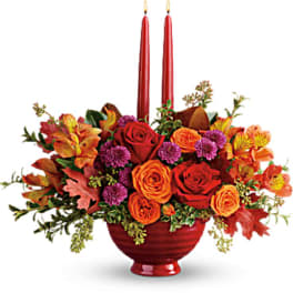 Autumn floral arrangement with red and orange roses in a red vase, accented by tall red candles.