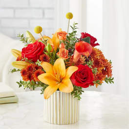 Mixed bouquet of red roses, yellow lilies, and orange flowers in a striped vase