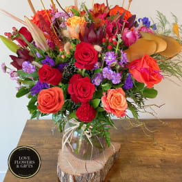 Tall mixed bouquet with red and orange roses, purple flowers, and cattails in a clear vase on a wood slice.